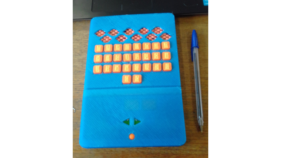A imagem apresenta um produto na cor azul e com tamanho e forma semelhantes a um pequeno livro. Ele está posicionado sobre uma mesa de madeira e ao lado de uma caneta esferográfica. Em destaque na face superior, encontram-se teclas retangulares na cor laranja e com celas Braille com identificação das letras do alfabeto
