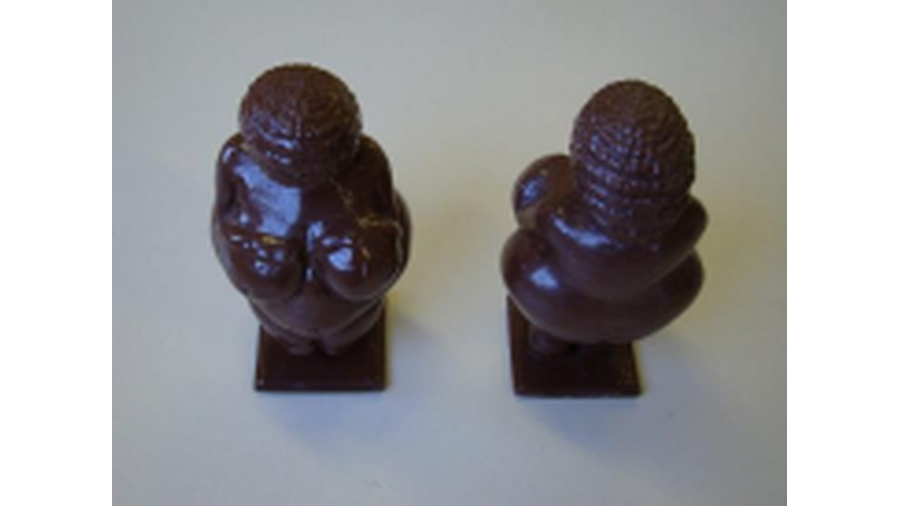 A imagem apresenta o recurso didático denominado Vênus de Willendorf, confeccionada em plástico por impressão 3D e retrata uma réplica em tamanho real da estatueta dessa obra, com as características dessa obra, uma figura feminina, seios e barriga extremamente volumosos, braços frágeis e imperceptíveis, tendo a cabeça coberta por algo que pode ser identificado como de rolos de tranças obstruindo sua face.