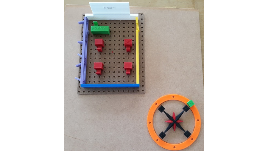 A imagem apresenta o recurso didático denominado Sala de Aula e Localização Espacial, que é composto por peças confeccionadas em impressão 3D, sendo elas as parede da sala de aula, as mesas dos alunos e professor e uma rosa dos ventos com identificação do norte e sul magnético e geográfico. As peças que representam a rosa dos ventos está montado sobre uma placa de MDF e as peças que compõem a sala de aula estão montados sobre uma placa perfurada e estão placa perfurada também esta montada sobre a placa de MDF. A rosa dos ventos e a sala de aula se orbitam na placa de MDF, conforme o usuário assim o fizer girar.
