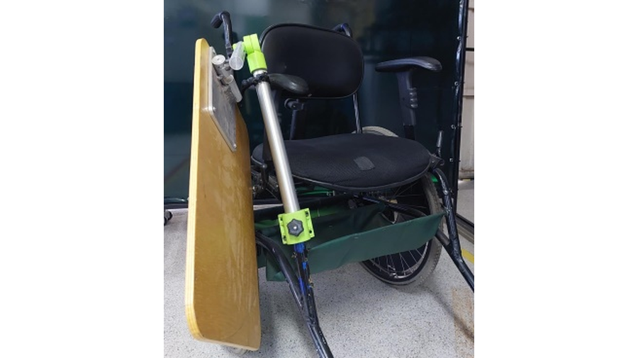 A imagem apresenta a foto de uma cadeira de rodas, com uma mesa, feita em braço em alumínio e base em madeira, acoplados a estrutura da cadeira. A mesa está posicionada lateralmente a cadeira.
