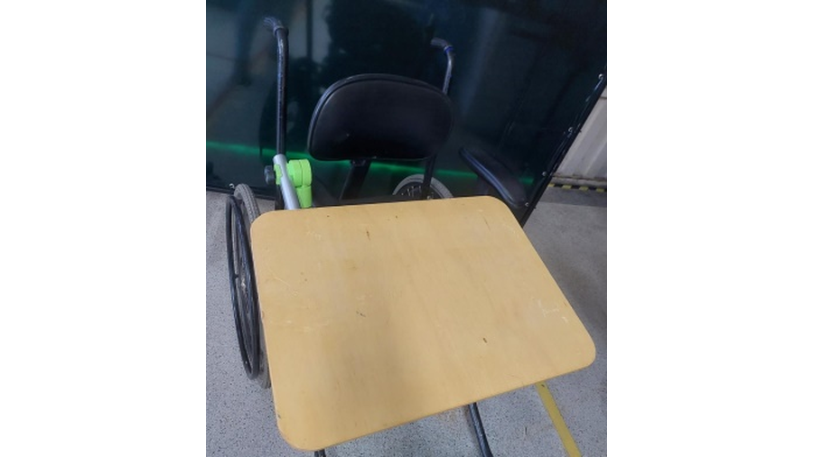 A imagem apresenta a foto de uma cadeira de rodas, com vista frontal, com uma mesa, feita em braço em alumínio e base em madeira, acoplados a estrutura da cadeira. A mesa está posicionada sobre a cadeira em condições de uso.