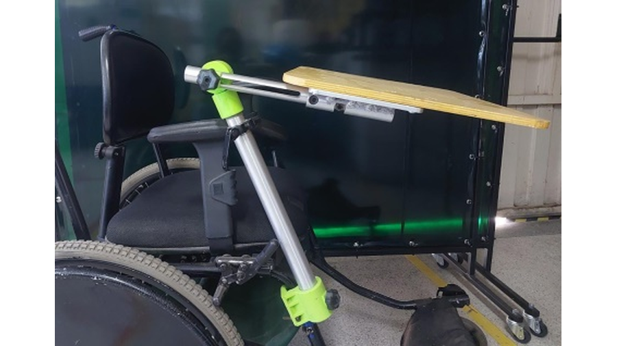 A imagem apresenta a foto de uma cadeira de rodas, com vista lateral, com uma mesa, feita em braço em alumínio e base em madeira, acoplados a estrutura da cadeira. A mesa está posicionada sobre a cadeira em condições de uso.