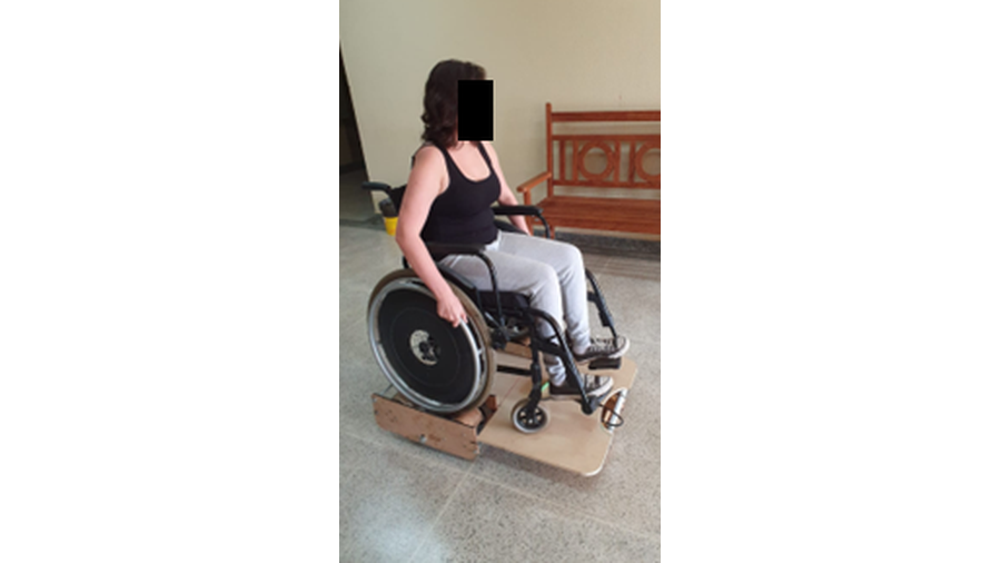 A imagem apresenta uma pessoa sentada numa cadeira de rodas, com as mãos sobre as rodas e posicionada sobre uma plataforma para elevação da cadeira de rodas. Essa plataforma é composta de chapas de MDF, quatro rodízios e dispositivos mecânicos para a realização do movimento conjuntamente com a cadeira.