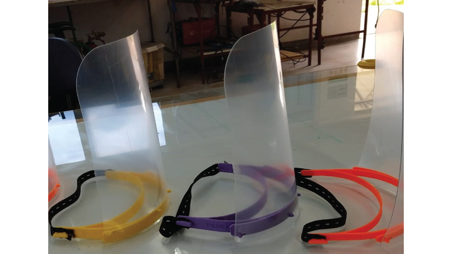 A imagem apresenta a fotografia de três protetores faciais Perfilados sobre uma mesa de tampo na cor branca. Cada protetor é constituído por uma folha transparente fixada a um arco plástico  colorido cujas extremidades encontram-se um segmento de elástico pretos com furos. As cores dos arcos são amarela, roxa e laranja.