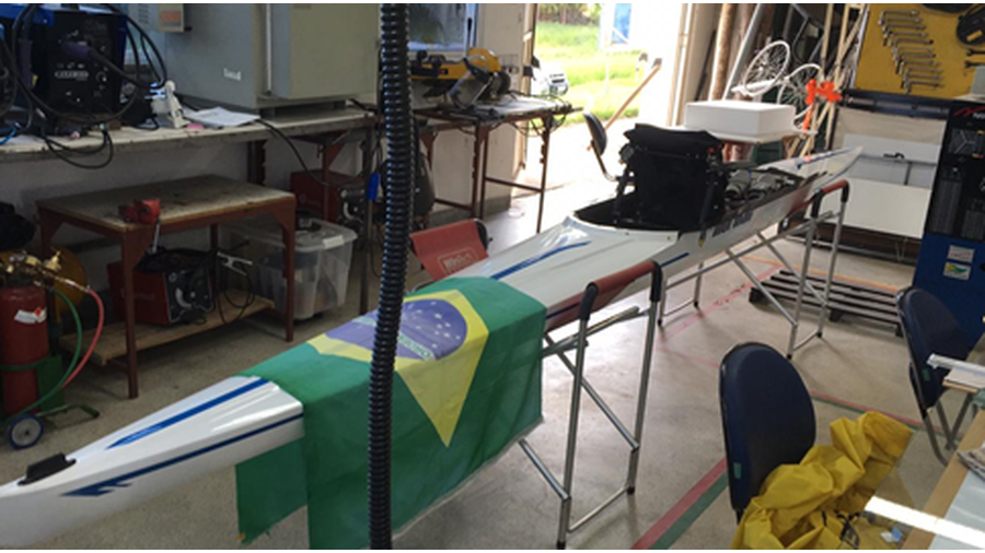 A imagem apresenta a foto de um barco a remo, apoiado sobre estruturas em alumínio, com uma bandeira do Brasil sobre o barco.